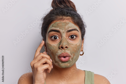 Portrait of astonished Indian woman with dark hair has shocked facial expression applies nourishing beauty mask on face to reduce blackheads and pores poses against white wall. Cosmetology treatment