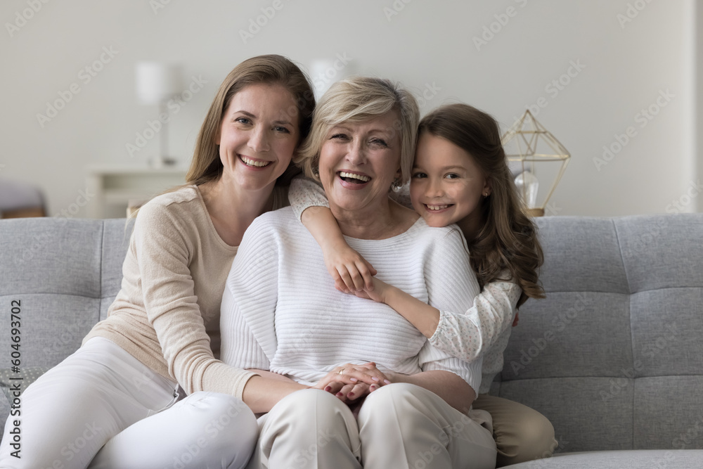 Three female generations family portrait. Cute little girl hugging tightly her beloved granny sit on sofa with young mom, smile look at camera, enjoy leisure time together. Unconditional love and ties Three female generations family portrait. Cute little girl hugging tightly her beloved granny sit on sofa with young mom, smile look at camera, enjoy leisure time together. Unconditional love and ties