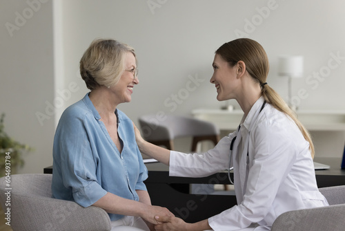Female doctor tells positive prognosis to aged woman patient, holds her hands, touches shoulder, provides support and medical care in hospital, take care of aged patient in private clinic. Healthcare photo