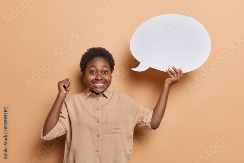 Studio shot of cheerful plump dark skinned young woman holds white blank speech bubble for your promotional content smiles broadly wears shirt and piercing in nose isolated over brown background.