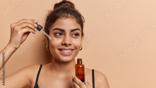 Domestic skin care and anti wrinkle routine. Pleased dark haired woman applies facial serum with dropper smiles toohthily focused aside dressed in t shirt isolated over brown background copy space