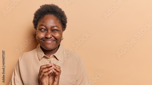 Fotografie Curious devious dark skinned woman steepls fingers and winks eye schemes somethi