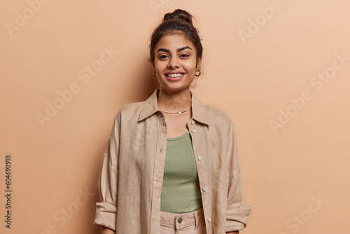 Horizontal shot of beautiful Indian woman smiles gently stands happy indoor wears shirt glad to hear pleasant words about her outlook poses against brown background. Smiling nice female model