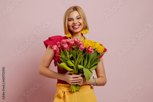 Beautiful young woman holding bunch of tulips and smiling against pink background