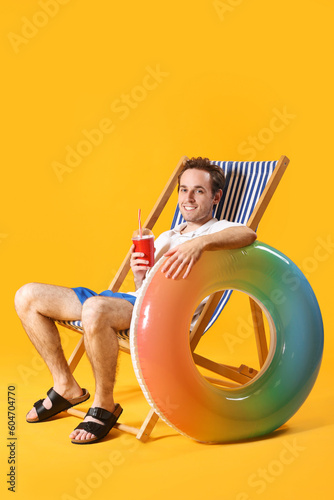 Young man with inflatable ring and soda sitting in deck chair on yellow background