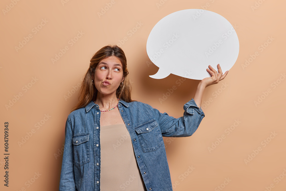 Thoughtful European woman with piercing in nose holds blank white speech bubble purses lips thinks what to write here dressed in denim shirt isolated over brown background. Let me think about it - obrazy, fototapety, plakaty 