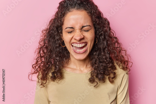 Photo of curly haired Brazilian woman keeps eyes closed exclaims loudly keeps mouth widwlt opened has white teeth dressed in casual brown jumper isolated over pink background. Human reactions concept