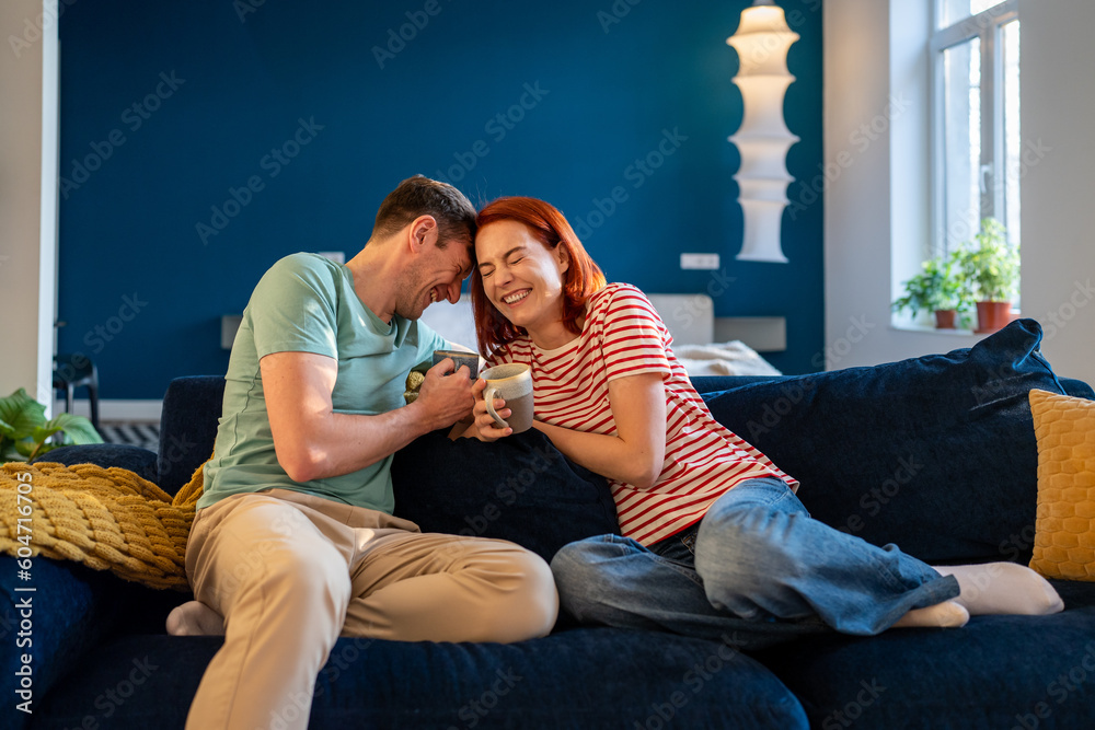 Happy carefree young couple in love joking laughing while relaxing on couch with coffee, bonding, enjoying morning time together at home. Emotional connection in a relationship