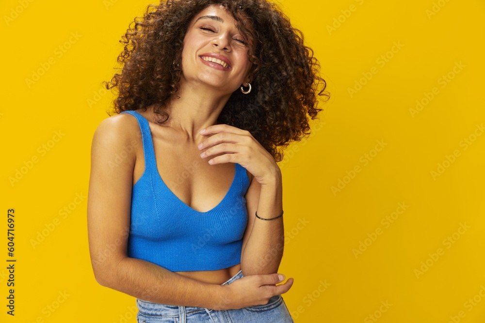 Woman with curly afro hair in a blue t-shirt on. yellow background signs with her hands, look into the camera, smile with teeth and happiness, copy space Woman with curly afro hair in a blue t-shirt on. yellow background signs with her hands, look into the camera, smile with teeth and happiness, copy space