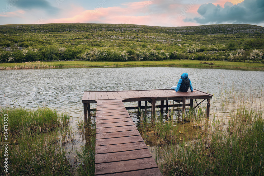 Hiker in blue jacket sitting on wooden pier by the lake with mountain in background at Burren national park, county Clare, Ireland, Adventure and lifestyle concept, wallpaper