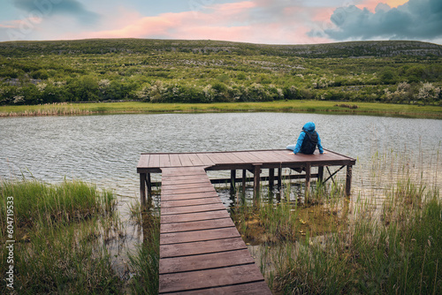 Obraz na płótnie Hiker in blue jacket sitting on wooden pier by the lake with mountain in background at Burren national park, county Clare, Ireland, Adventure and lifestyle concept, wallpaper w salonie