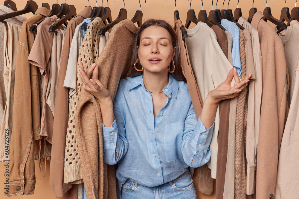Horizontal shot of European woman meditates among clothes rail keeps eyes closed dressed in shirt and jeans tries to find inner balance before choosing outfit to wear. Yoga practice near wardrobe