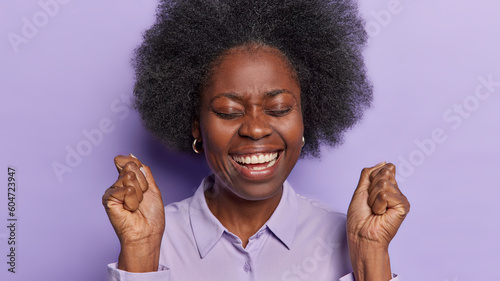 Charming optimistic African woman feels joyful smiles toothily shakesclenched fists keeps eyes closed and triumphs over something got success on work isolated on purple background. Yeah finally I won