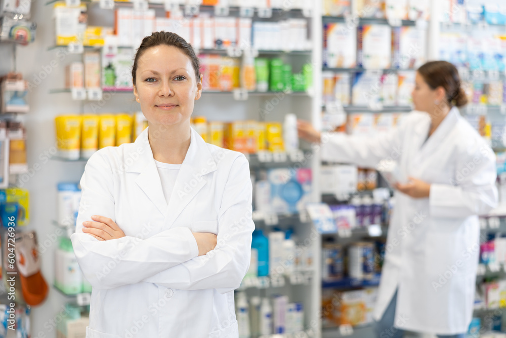 Adult female pharmacist in uniform posing against backdrop of shelves with products in pharmacy