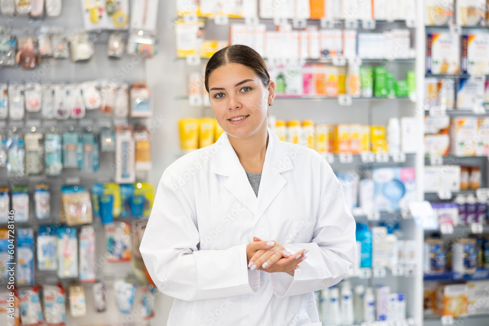 In pharmacy, positive female chemist stands and waits for visitors. Wide range of prescription and over-counter medications, patient care products, baby food