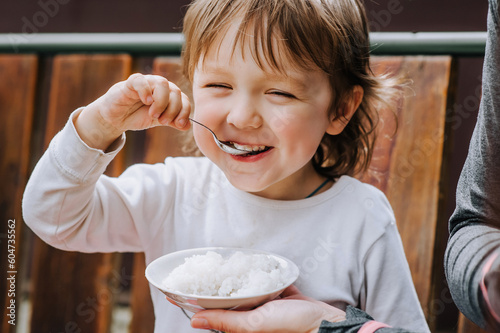 Handsome long-haired little boy of preschool age, happy child eats rice porridge with a spoon in his hand with the help of his mother. Photography, close-up portrait, baby food, feeding, concept.