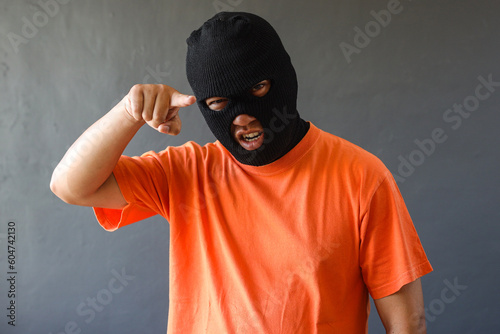 Фотография Prisoner in orange shirt and black mask pointing finger to camera over grey wall