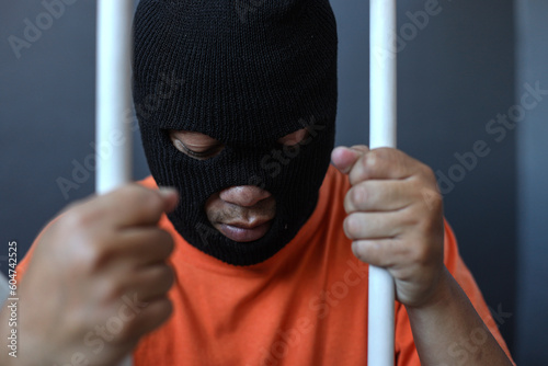 Obraz na plátně a prisoner in orange shirt and black mask inside the bars of a prison