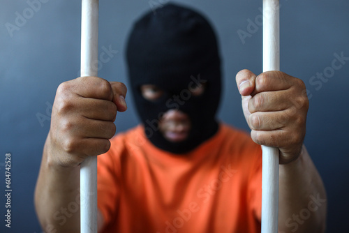 a prisoner in orange shirt and black mask inside the bars of a prison 