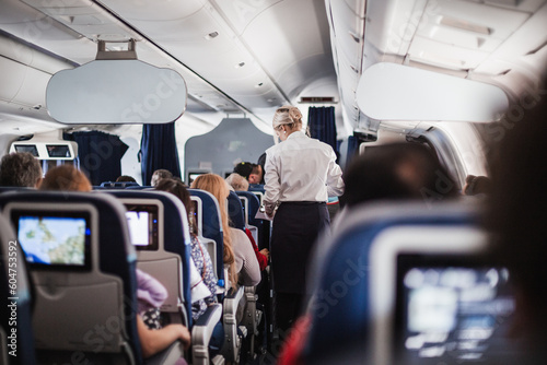 Interior of airplane with passengers on seats and stewardess in uniform walking the aisle, serving people. Commercial economy flight service concept