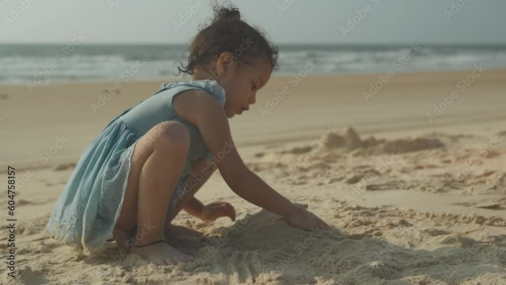 The girl sits on the sandy shore and builds a sand castle - Happy little girl plays with sand on the beach in the background at the blue sea at sunset.