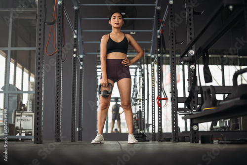 Strong Asian woman doing exercise with kettlebell at cross fit gym. Athlete female wearing sportswear workout on grey gym background with weight and dumbbell equipment. Healthy lifestyle.