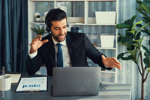 Φωτογραφία Male call center operator or telesales representative siting at his office desk wearing headset and in conversation with client providing customer service support or making persuasive sale