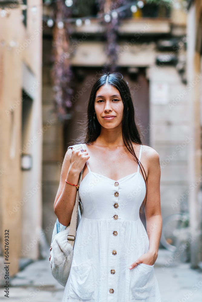 Hispanic woman with backpack holding hand in pocket of white dress and looking at camera while standing on blurred background of street of Barcelona, Spain