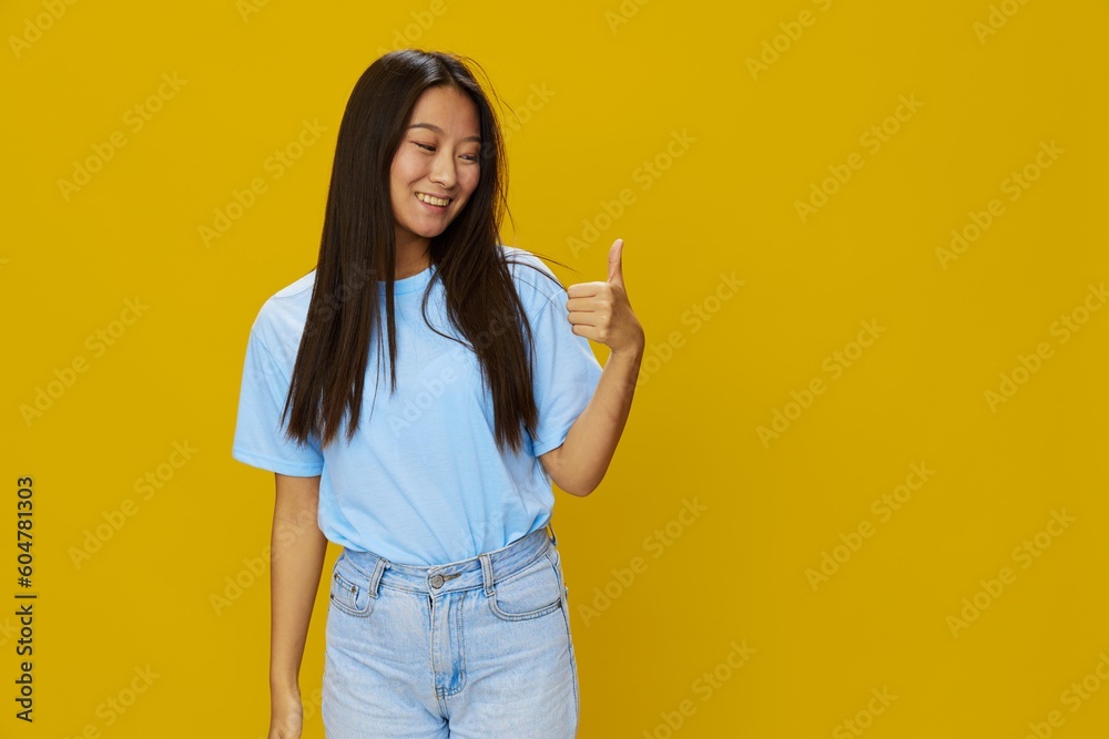 Asian woman smile with teeth hand gestures, signs and symbols on yellow background in blue t-shirt and jeans Asian woman smile with teeth hand gestures, signs and symbols on yellow background in blue t-shirt and jeans