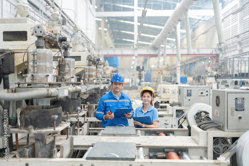 American engineers and Asian men and women, smart looking, standing with confidence, holding list notes, in plastic industry factory. There is a working machine Wear uniforms and helmets.