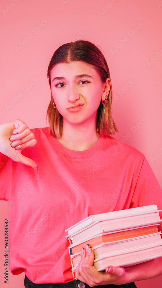 Dislike student. Boring education. Exam failure. Dissatisfied sad girl holding stack of books showing thumb down sign grimacing isolated on neon color pink background. Dislike student. Boring education. Exam failure. Dissatisfied sad girl holding stack of books showing thumb down sign grimacing isolated on neon color pink background.