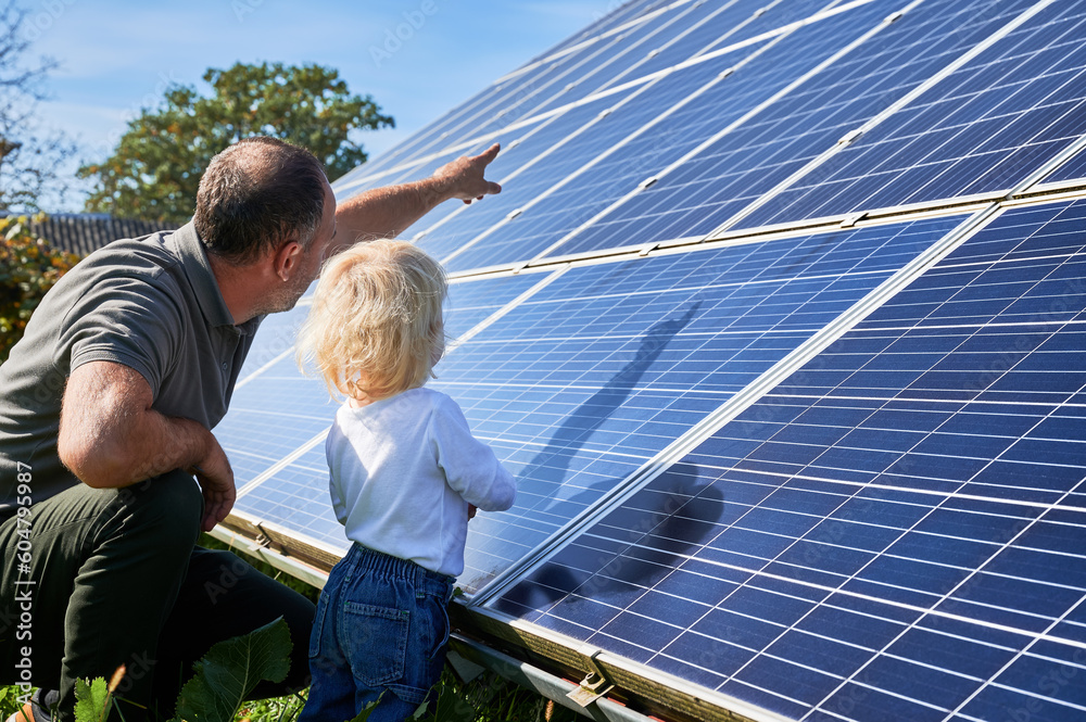 Man showing his small child the solar panels during sunny day. Father presenting to son modern energy resource. Back view of father and his kid watching on solar panels - obrazy, fototapety, plakaty 