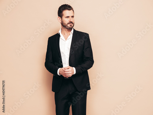 Portrait of handsome confident stylish hipster lambersexual model. Sexy modern man dressed in black elegant suit. Fashion male posing in studio, isolated on beige