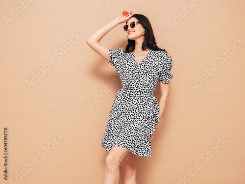 Young beautiful smiling female in trendy summer dress. Sexy carefree woman posing in studio on beige background. Positive brunette model having fun. Cheerful and happy. Isolated. In sunglasses