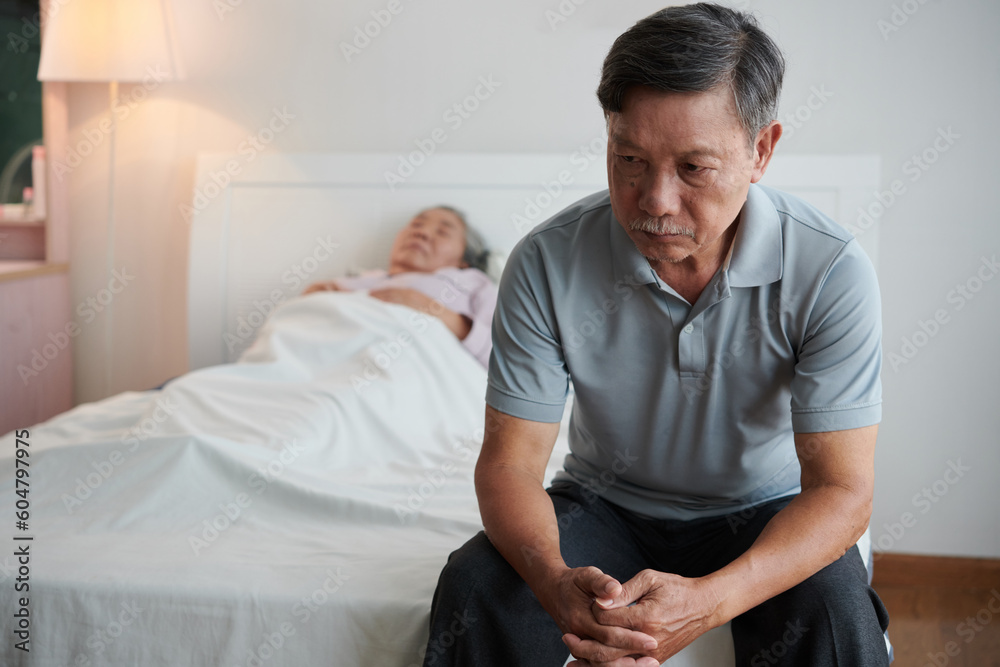 Fototapeta premium Depressed senior man deep in thoughts sitting on bed of his sick wife