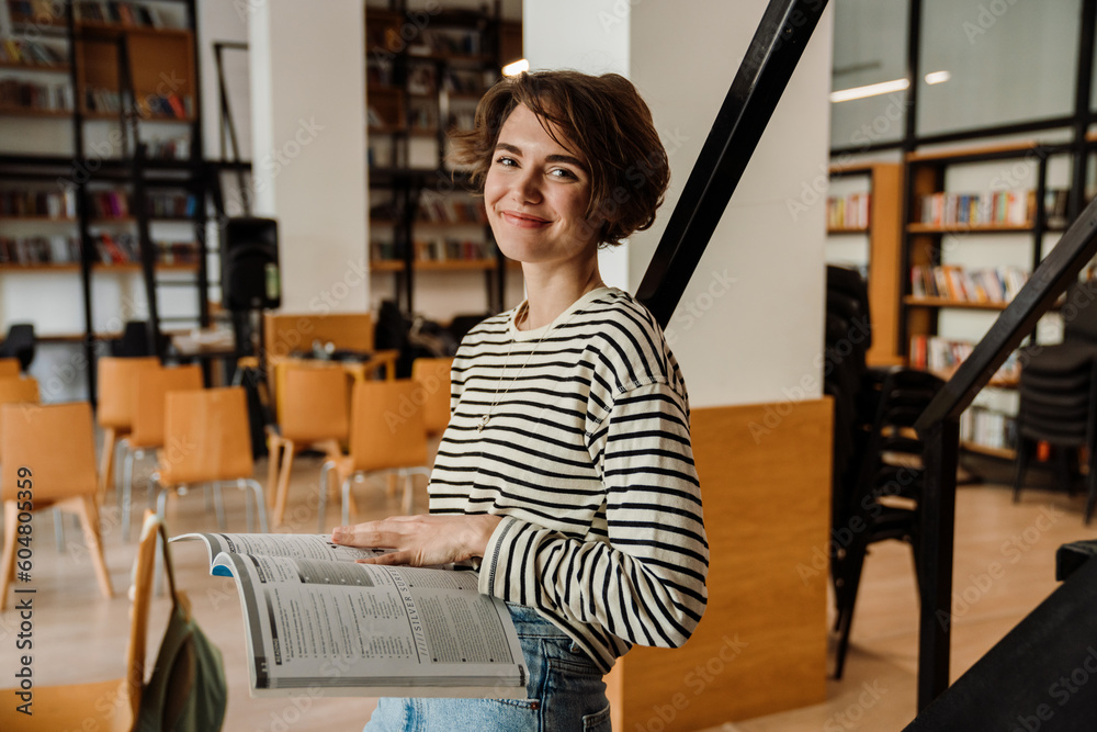 Smiling woman student holding textbook while standing in library Smiling woman student holding textbook while standing in library
