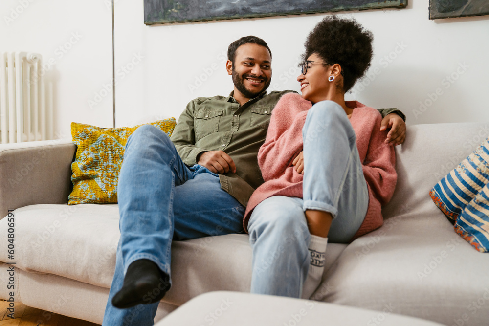 Cheerful multiethnic couple hugging while sitting on couch at home Cheerful multiethnic couple hugging while sitting on couch at home