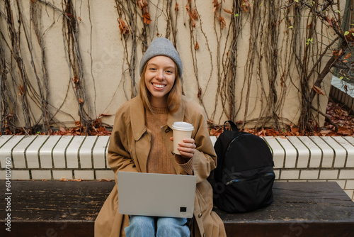 Cheerful girl working on laptop and drinking coffee while sitting on bench outdoors