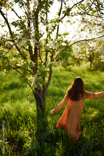 a beautiful, joyful woman is standing in a long orange dress, in the countryside, near a tree blooming with white flowers, during sunset, illuminated from the back and joyfully spinning