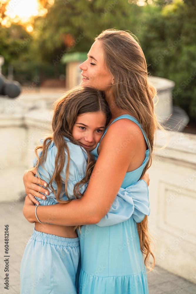 Fototapeta premium Mother daughter sunset. in blue dresses with flowing long hair against the backdrop of sunset. The woman hugs and presses the girl to her.