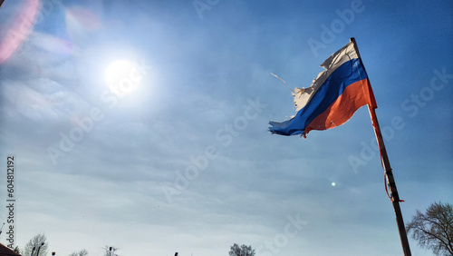 A tattered flag of Russia against the backdrop of a blue sky with the sun. The confrontation between Russia and Ukraine and the military actions of the countries with victories and defeats