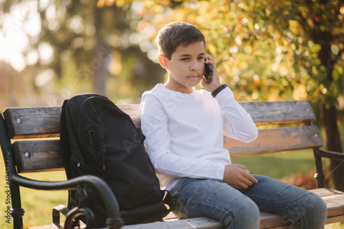 Young boy in white sweatshirt with black backpack sitting on the bench in the park and speak with somebody by the phone