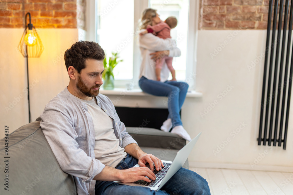 Fototapeta premium Bearded man working on computer, wife and toddler daughter on the background hugging.