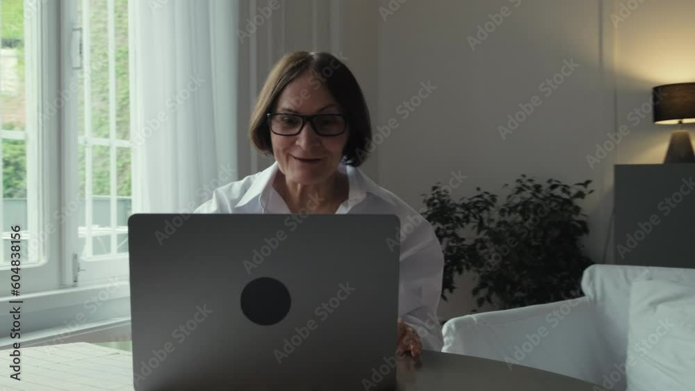 An elderly woman with dark hair in a white shirt sits at a round wooden table in front of a laptop and communicates via video link. Video call. Communication of people in the network. An elderly woman with dark hair in a white shirt sits at a round wooden table in front of a laptop and communicates via video link. Video call. Communication of people in the network.
