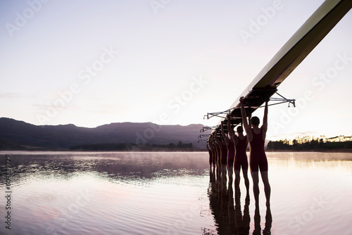 Rowing team entering lake at dawn with scull overhead 