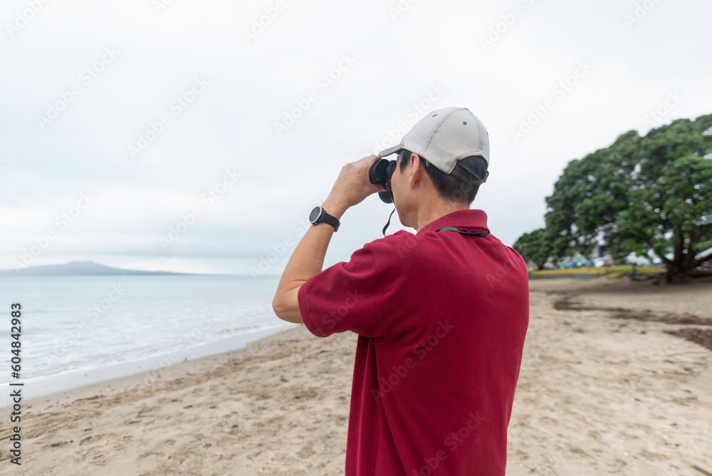Obraz premium Man holding binoculars looking at Rangitoto Island. Milford Beach. Auckland.