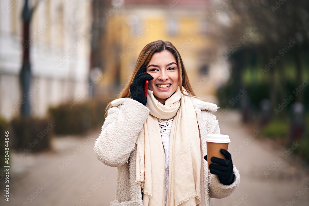 Fototapeta premium Young cheerful woman walking on city streets with hot coffe in her hand while talking on her mobile phone - Outdoor smartphone usage concept in cold winter weather with a girl wearing jacket