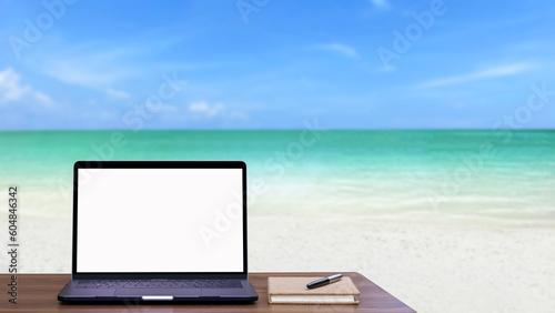 Mockup laptop computer with blank white screen and notebook on wooden table with sea beach view blur background. Summer and vacation concept.