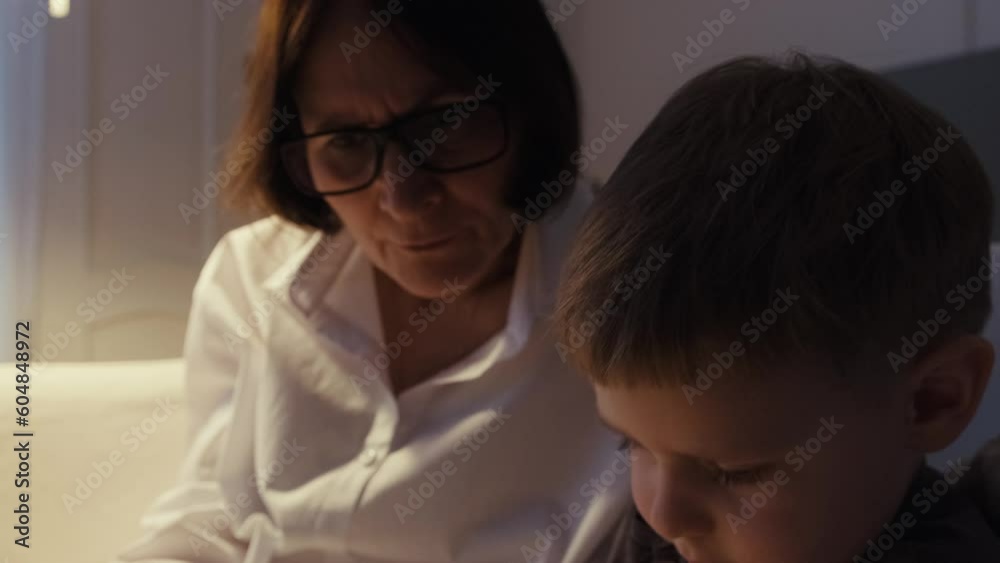 Grandmother and grandson are sitting on a white sofa and reading a book together. A cozy evening at Grandma's. Senior lady and her grandson spend time together. Grandmother and grandson are sitting on a white sofa and reading a book together. A cozy evening at Grandma's. Senior lady and her grandson spend time together.