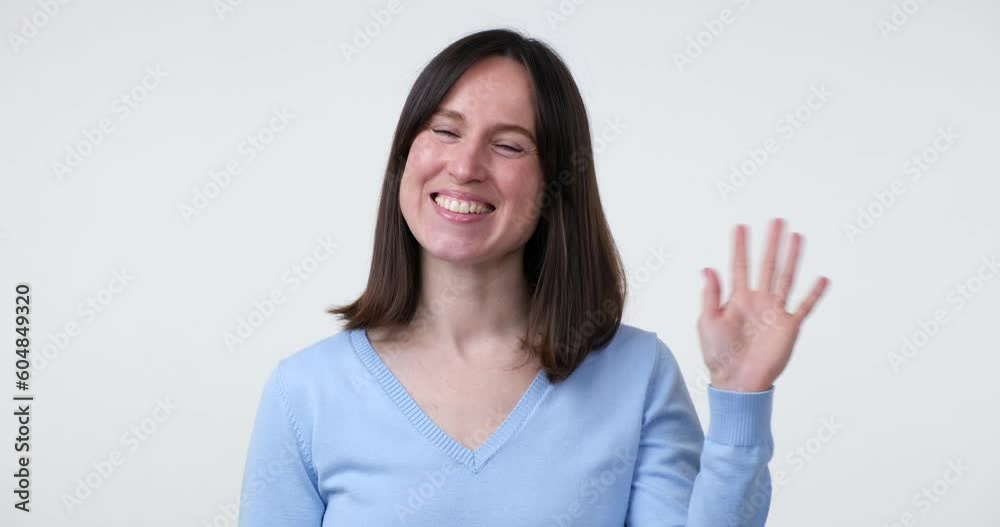 A cheerful Caucasian woman standing on a white background waves her hand in a welcoming gesture and flashes a warm smile, expressing her friendliness and good mood. A cheerful Caucasian woman standing on a white background waves her hand in a welcoming gesture and flashes a warm smile, expressing her friendliness and good mood.